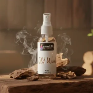 A spray bottle of "Old Wood" fragrance with a white label, centered on a rustic wooden block. Around the bottle are pieces of dark, textured oud wood and wisps of white smoke, creating an earthy and woody ambiance. In the background, there are stacked logs and some green leaves, enhancing the natural, forest-like theme.