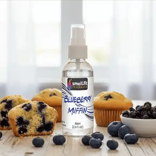 Product image of Smell.fit Fragrance 'Blueberry Muffin' 80ml (2.8 FL OZ) mist spray bottle. The bottle is surrounded by fresh blueberries, several baked blueberry muffins, and a small white bowl filled with dried blueberries, set on a rustic wooden table.