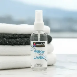 Product image of Smell.fit Fragrance 'Fresh Linen' 80ml (2.8 FL OZ) mist spray bottle. The bottle is positioned in front of a neat stack of clean, folded white and dark gray towels, suggesting a laundry or bathroom setting.