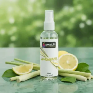Product image of Smell.fit Fragrance 'Lemongrass' 80ml (2.8 FL OZ) mist spray bottle. The bottle is surrounded by fresh stalks of lemongrass, bright yellow lemon slices, and small green leaves, set on a glossy green surface against a blurred, leafy green background.