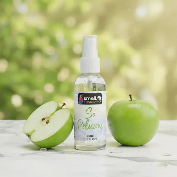 Product image of Smell.fit Fragrance 'So Delicious' 80ml (2.9 FL OZ) mist spray bottle. The bottle is placed on a white marble surface with water droplets, flanked by a whole green apple and a perfectly sliced half of a green apple, against a blurred, sunny green outdoor background.