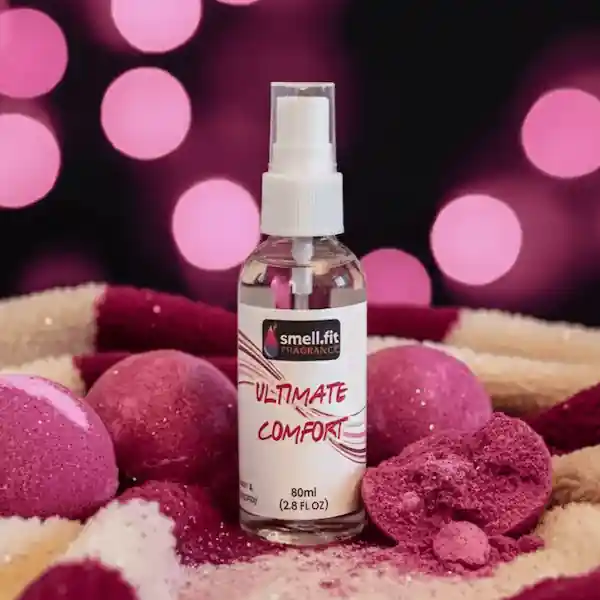 Product image of Smell.fit Fragrance 'Ultimate Comfort Room & Bodyspray' 80ml (2.8 FL OZ) mist spray bottle. The bottle is set on a soft, cozy surface surrounded by sparkly pink bath bombs (some whole, some broken or crumbled), against a blurred background of warm pink bokeh lights.